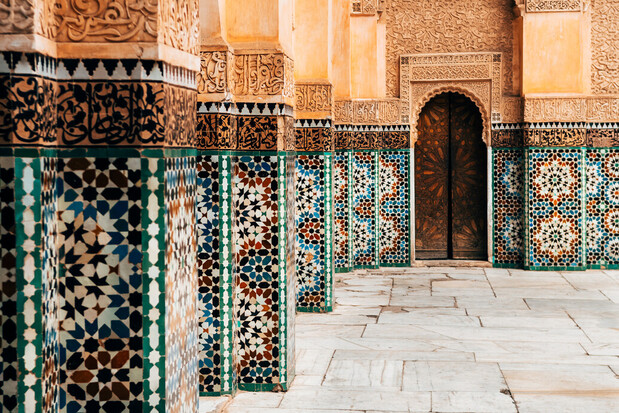Ornate columns feature intricate geometric tile patterns, leading to a carved wooden door in a richly decorated architectural setting with detailed relief work. The floor consists of large stone tiles.
