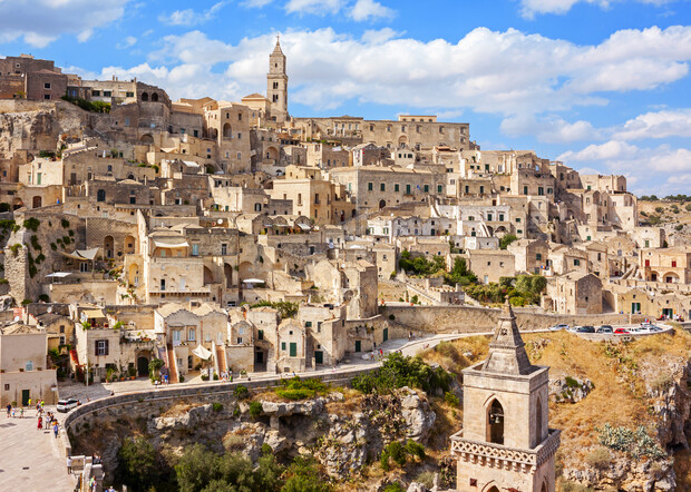 Ancient stone buildings form a hillside town under a blue sky with scattered clouds. A narrow road curves through the terraced structures, featuring a prominent bell tower in the foreground.