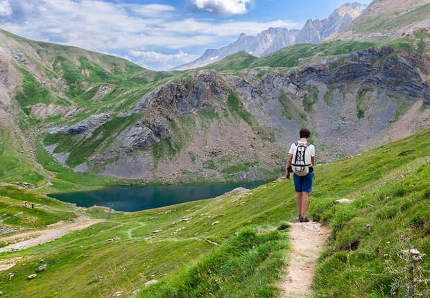 Hiker walks along a narrow, grassy trail overlooking a mountainous landscape with a small, tranquil lake nestled in a green valley under a partly cloudy sky.
