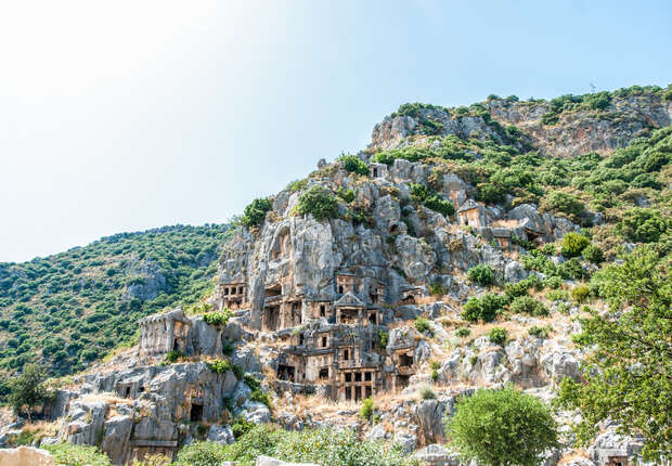 Rock-cut tombs are carved into a steep cliff, surrounded by greenery and under a clear blue sky, reflecting an ancient architectural style embedded within a natural landscape.
