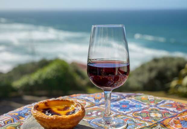 A glass of red wine sits on a colorful tiled table beside a golden pastry, with an ocean view in the background under a clear sky.