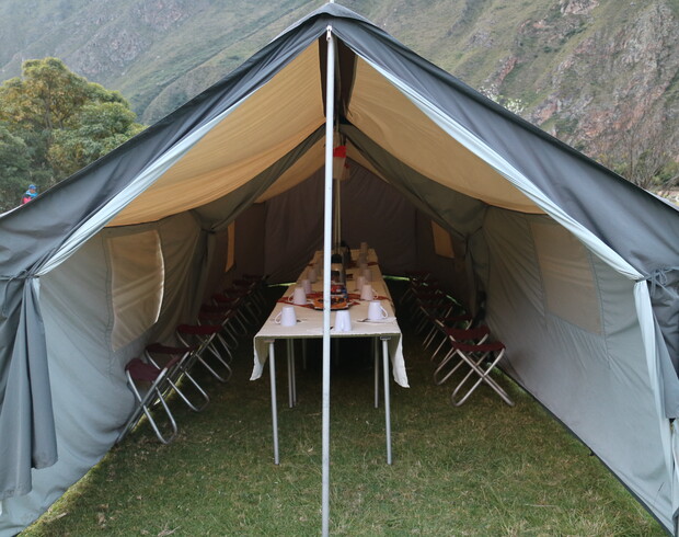 A large open tent houses a long table set with cups and dishes, surrounded by folding chairs. It is situated on a grassy area with distant mountains.