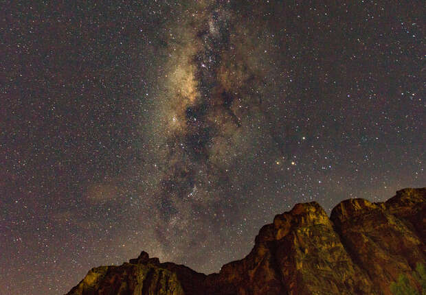 Starry sky showcases the Milky Way stretching vertically, with rocky cliffs silhouetted against the night, creating a dramatic contrast between the illuminated galaxy and the dark landscape below.