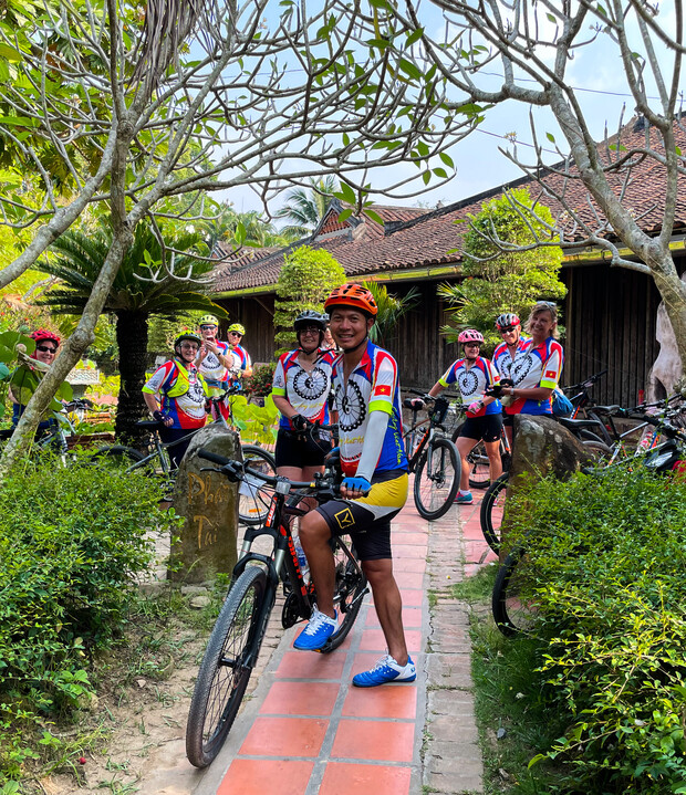 Cyclists in colorful jerseys and helmets pose with bikes on a tiled path, surrounded by lush greenery and trees, in front of a traditional wooden building.