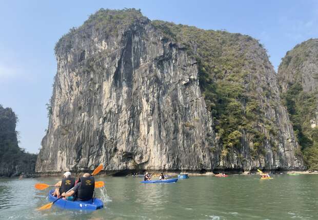 Kayakers paddle on turquoise water, surrounded by towering limestone cliffs with lush greenery. Several boats are visible in the peaceful, natural setting under a clear blue sky.
