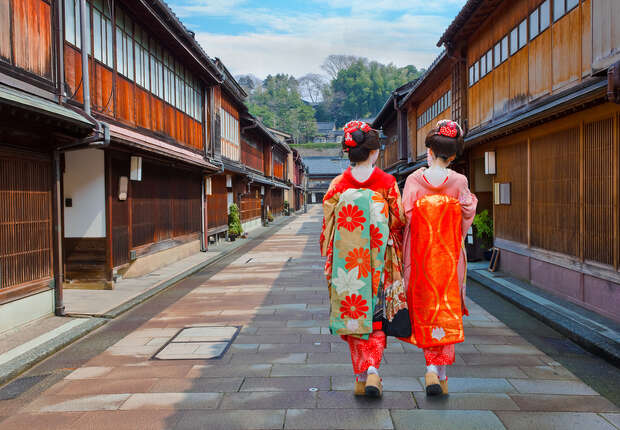 Two people dressed in colorful traditional kimonos walk down a narrow, paved street, bordered by wooden buildings, with trees visible in the distance.