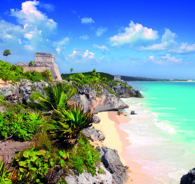 Ruins rest atop a rocky cliff overlooking a turquoise sea and sandy beach, surrounded by lush vegetation and palm trees, beneath a bright blue sky with scattered clouds.