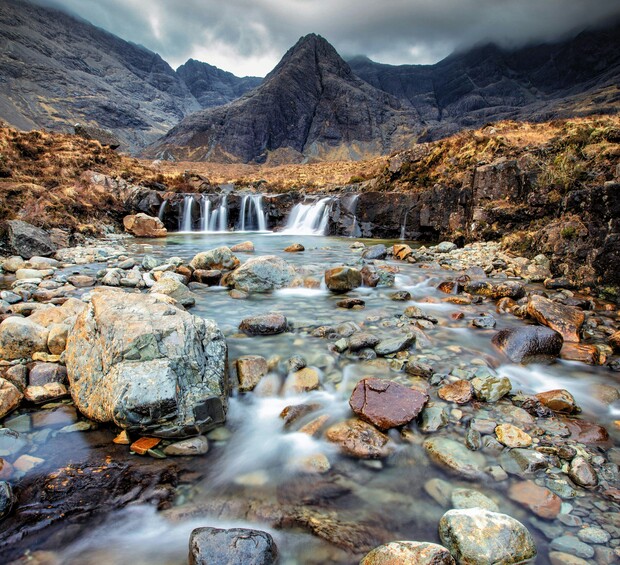 A mountain stream cascades over rocks into small waterfalls, surrounded by rugged terrain, with misty mountains towering in the background under a cloudy sky.