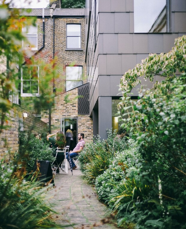 A group of people sit and converse at a patio table nestled between brick buildings. Lush greenery surrounds the path leading to the gathering.