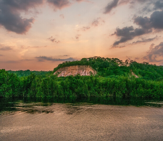Clouds drift over a lush, green hillside with a rocky cliff, bordered by thick vegetation. A calm river flows in the foreground, at sunset, with a vibrant, colorful sky.