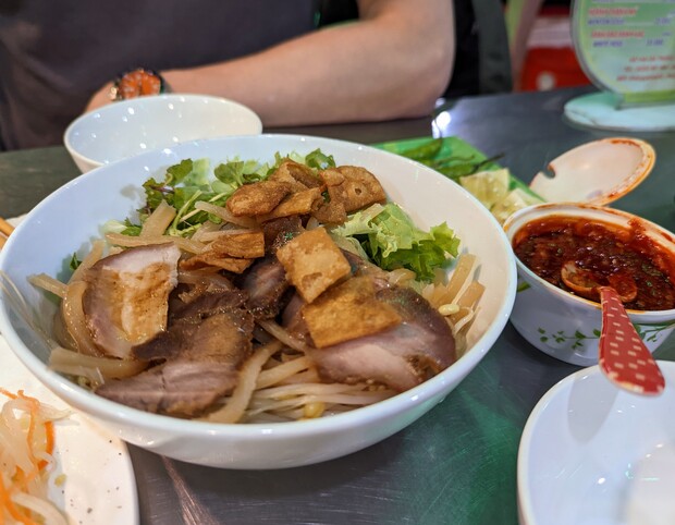 A bowl of noodles topped with sliced meat, greens, and crispy topping is placed on a metal table. Nearby, there is a small bowl of red sauce with a spoon.