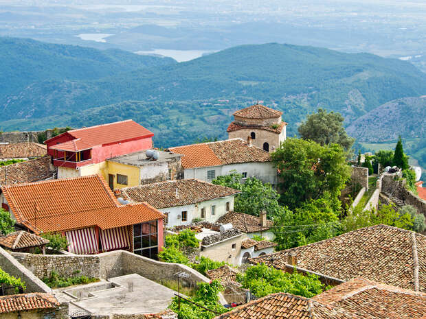 Tile-roofed houses nestled on a hillside overlook expansive, lush mountains and a distant calm body of water under a clear sky. A mix of greenery and stone walls surrounds the village.