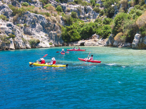 Kayakers paddle colorful kayaks across vibrant blue water in a rocky cove surrounded by lush greenery and rugged cliffs under a clear sky.