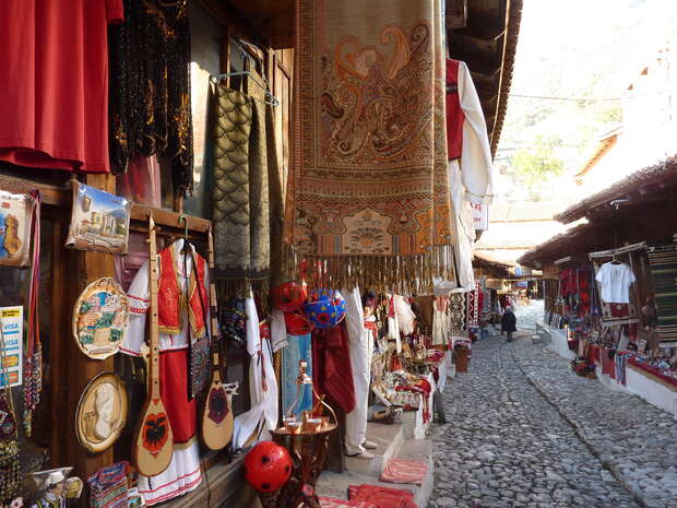 Textile market stalls display vibrant rugs, clothing, and handicrafts, lining a narrow cobblestone street. Sunlight filters through, casting shadows on colorful goods, creating a bustling and lively marketplace atmosphere.