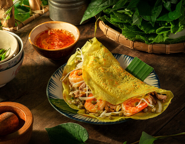 A yellow pancake with shrimp, bean sprouts, and meat sits on a blue-white plate, accompanied by a bowl of orange dipping sauce, surrounded by fresh greens on a wooden table.