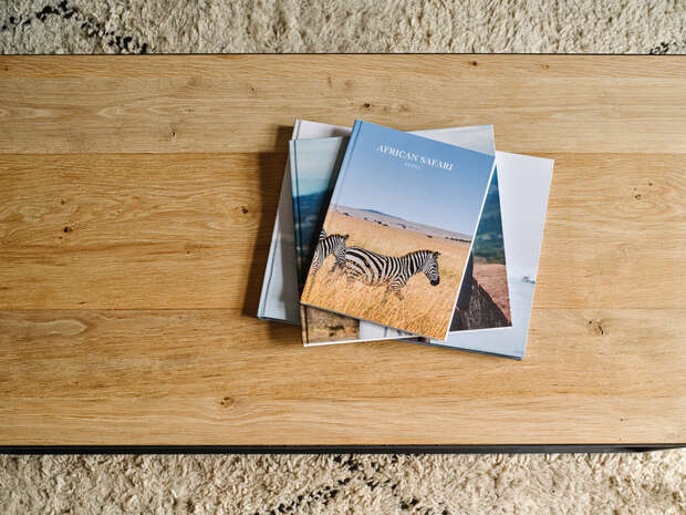 Books lie stacked on a wooden table, with the top book titled "AFRICAN SAFARI" featuring a zebra in a grassy landscape on its cover, situated in a carpeted room.