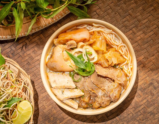 A bowl of noodle soup with sliced meat and herbs sits on a woven mat, garnished with lime and fresh greens in a rustic setting.