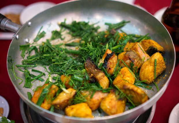 Chunks of cooked fish seasoned with vibrant green herbs are arranged in a silver pan, surrounded by a red tablecloth and white dishes, suggesting a meal setting.
