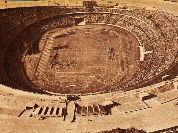 A large, oval stadium filled with spectators surrounds a grassy field, featuring a track. The atmosphere suggests an event, with structures and seating arranged around the perimeter.