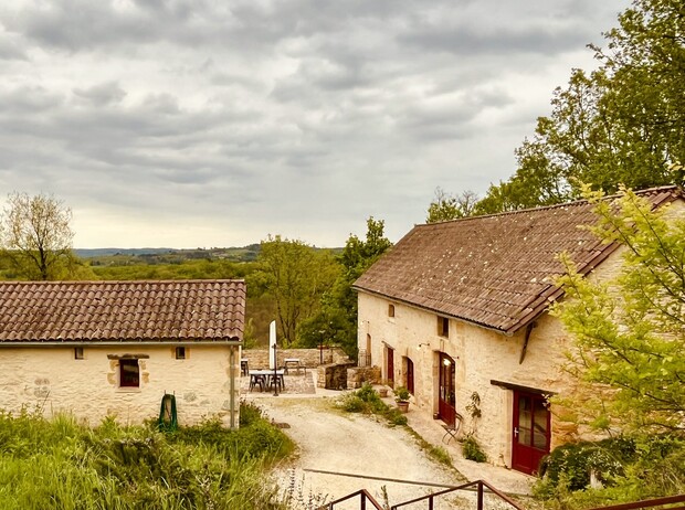 Stone cottages with red doors stand quietly, surrounded by lush greenery and under a cloudy sky, set in a rural landscape overlooking distant rolling hills.