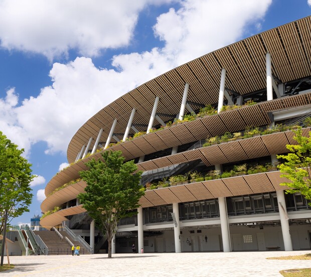 A modern stadium with tiered, plant-filled terraces under a bright blue sky. Tall vertical beams support the structure, surrounded by trees and a few people walking nearby.