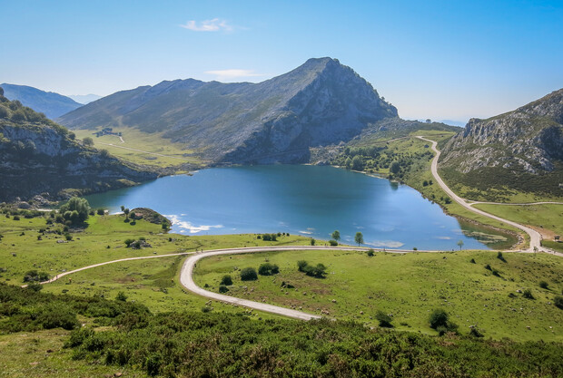 A serene, reflective lake nestled among rolling green fields and rugged mountains, under a clear blue sky. A winding road curves through the landscape, enhancing the tranquil scenery.