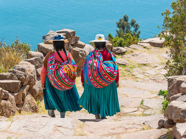 Two women wearing traditional colorful clothing and hats walk down a stone path, carrying striped shawls on their backs. They are surrounded by greenery, overlooking a blue water body.