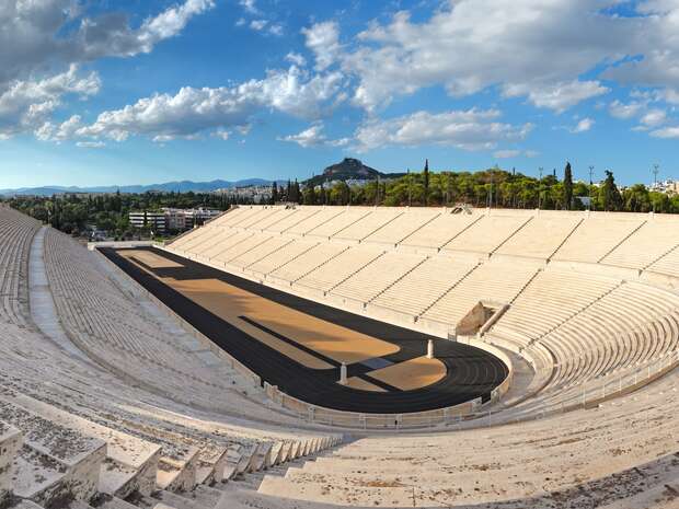 A historic marble stadium sits empty, with a black track encircling a sandy field. Hilltops and trees surround the structure under a partly cloudy blue sky.