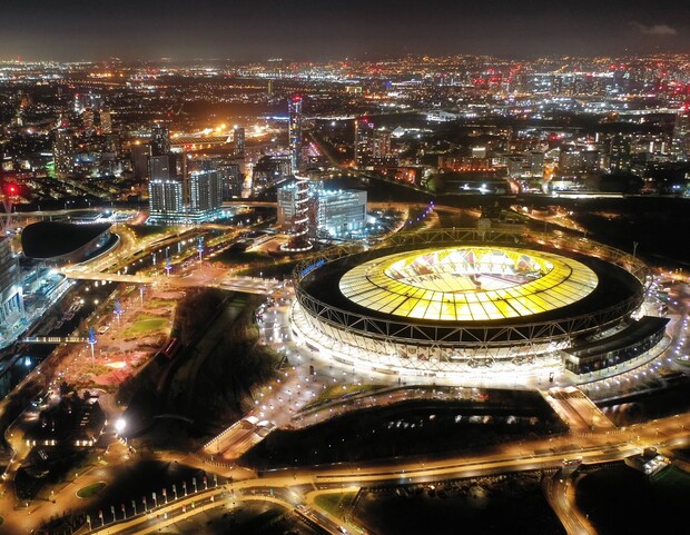A brightly lit stadium glows amidst a sprawling urban area at night, surrounded by illuminated buildings and roads, creating a vibrant cityscape.
