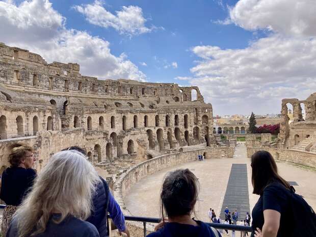 Ancient amphitheater stands in partial ruins under a cloudy sky, with visitors observing from a balcony, providing a sense of historical exploration and architectural grandeur.