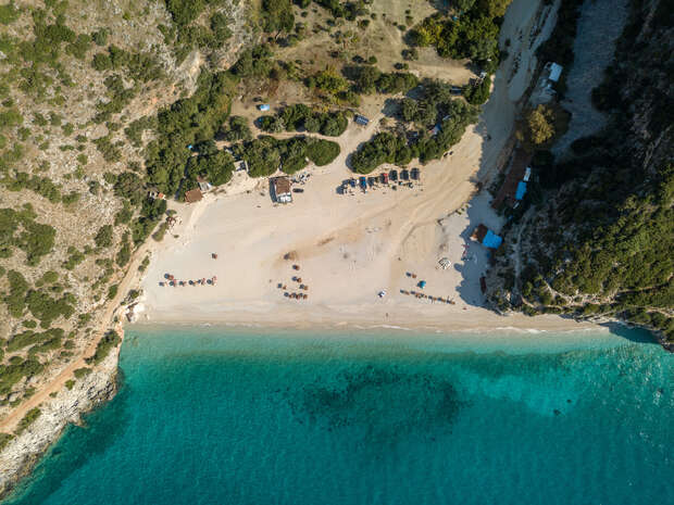 Beach bordered by turquoise water and rugged cliffs, with scattered umbrellas and sunbeds. Trees and small structures dot the sandy shoreline, creating a serene, secluded cove ambiance.