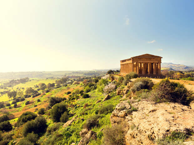 An ancient Greek temple with tall columns stands on a rocky hill, overlooking a vast, verdant landscape under a clear sky.