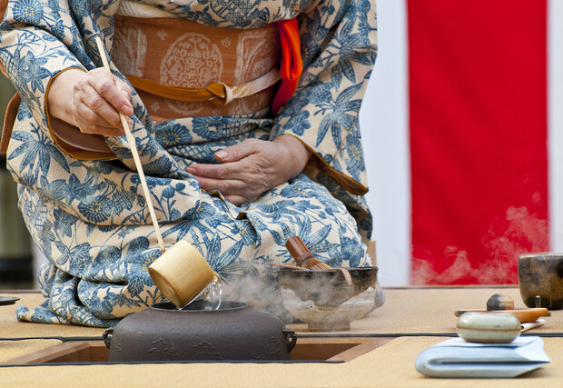 A person in a floral-patterned kimono uses a bamboo ladle to pour hot water from an iron kettle during a tea ceremony. The setting includes bowls and a paper mat.