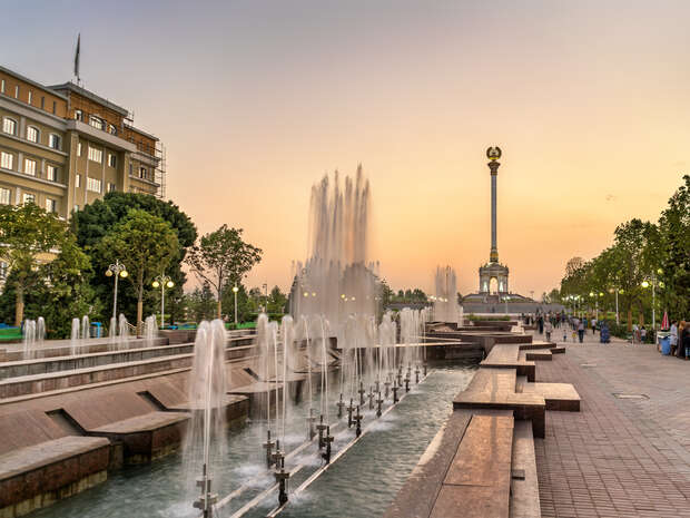 Fountain jets rise from a long, rectangular pool in a park; pedestrians stroll along the paved path. A tall monument with a statue is visible against the sunset sky.