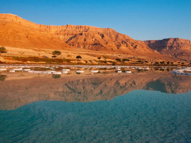 Calm water reflects rugged, golden-brown mountains under a clear blue sky, with white salt formations around the water's edge, creating a serene, natural landscape contrasting with barren terrain.