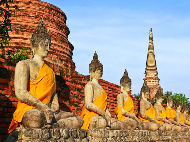 Stone Buddha statues, draped in orange robes, sit serenely in a lined row against a backdrop of ancient red brick ruins and a tall spire under a clear blue sky.