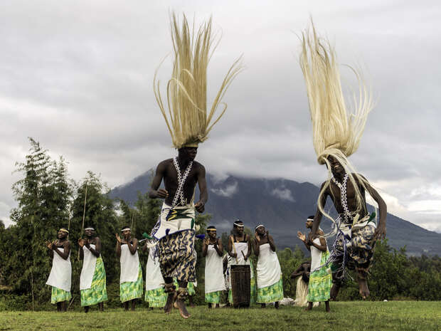 Dancers leap energetically, their blond headdresses soaring, as musicians play drums and wind instruments. The vibrant traditional costumes enhance the scene against a backdrop of lush greenery and distant mountains.