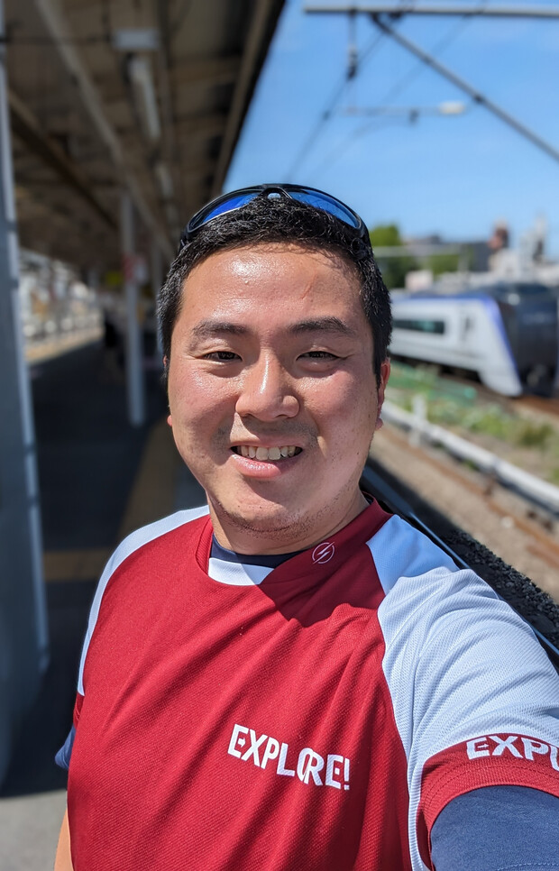 A person wearing a red and white shirt with "EXPLORE!" written on it smiles while taking a selfie at a train station platform, with a blurred train passing in the background.