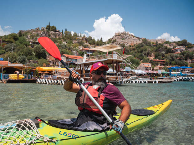 A person wearing a red hat and life vest paddles a yellow kayak in clear water, surrounded by a scenic coastal village with hills and structures under a partly cloudy sky.