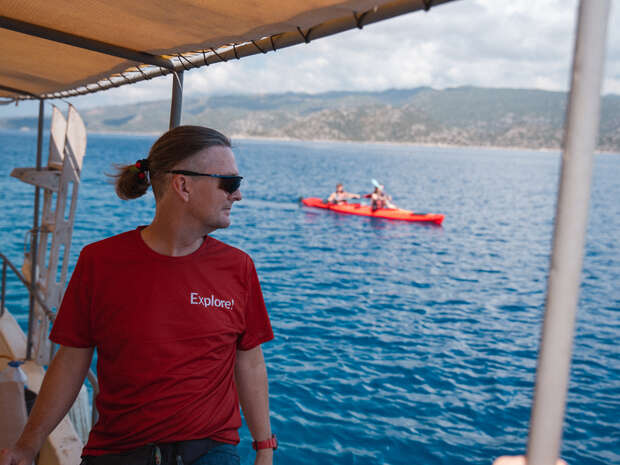 A person wearing a red "Explore!" shirt and sunglasses stands on a boat looking at a kayak in the blue sea near distant mountains under a partially cloudy sky.