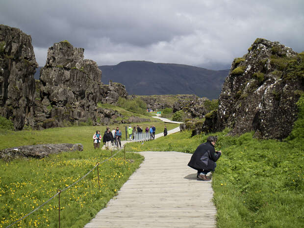 A pathway winds between rocky cliffs and green grass. A person crouches to photograph flora while others walk in the distance. Mountains and overcast skies complete the scenic landscape.