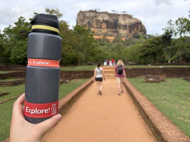 A hand holds a gray water bottle labeled "Explore!" on a path leading toward a large rock formation. In the background, two people walk along the path surrounded by trees.