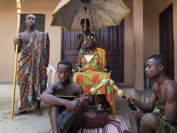 A person in ceremonial attire sits on a throne under an umbrella. Two individuals kneel, playing traditional instruments, while another stands holding a staff, all against a backdrop of a building.