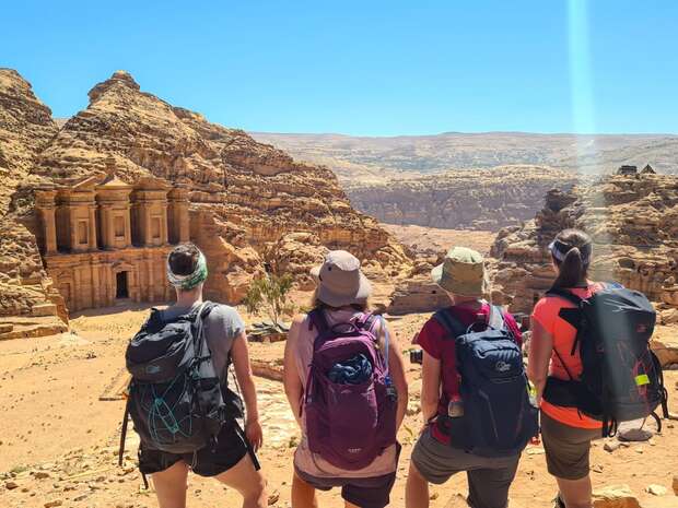 Four hikers with backpacks stand facing an ancient rock-carved building, resembling Petra's architecture, in a rugged desert landscape under a clear blue sky.