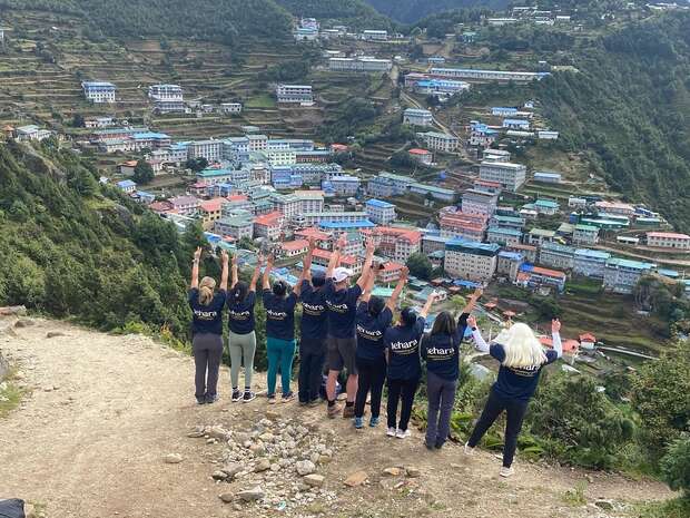 A group of eight people stand on a rocky path with arms raised, overlooking a hillside village with colorful buildings. Their shirts display the text: “klara”. Lush greenery surrounds the area.