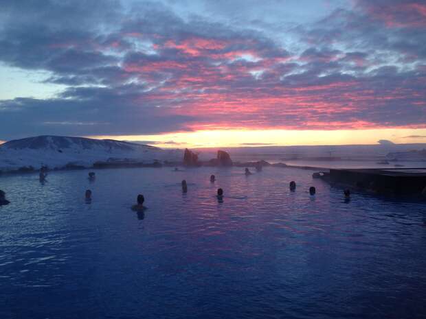 Silhouetted people relax in a large outdoor pool. The water reflects a vibrant pink and purple sunset, contrasting against a snowy landscape and a dramatic, cloudy sky in the background.