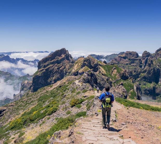 A hiker wearing a backpack walks down a stone path through a mountainous landscape, surrounded by green vegetation and low clouds beneath a clear blue sky.