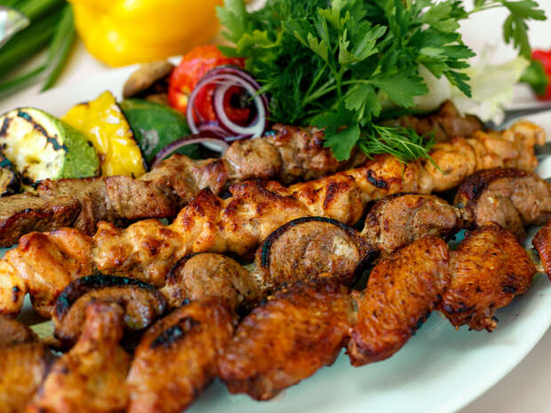 Grilled skewers featuring seasoned meats are displayed on a plate, surrounded by roasted vegetables and fresh herbs, including parsley and dill, against a backdrop of various colorful vegetables.