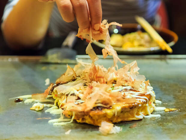 A hand sprinkles bonito flakes onto sizzling okonomiyaki on a hot griddle, with a background showing a plate and chopsticks.