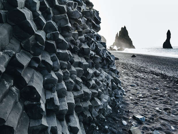 Hexagonal basalt columns rise sharply along the shoreline, with waves crashing in the distance and sea stacks protruding from the water under a misty sky.